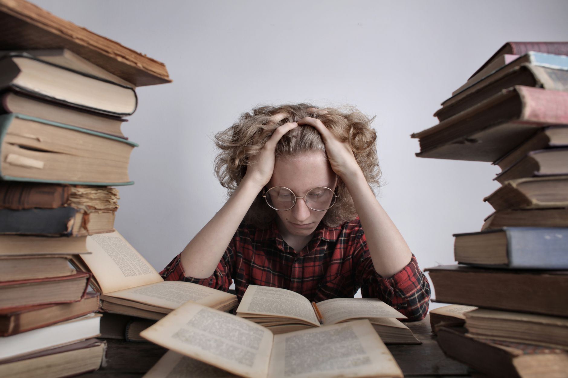 Tired teenage boy in casual clothes and eyeglasses sitting at desk with large stacks of books holding head and reading book while preparing for exam