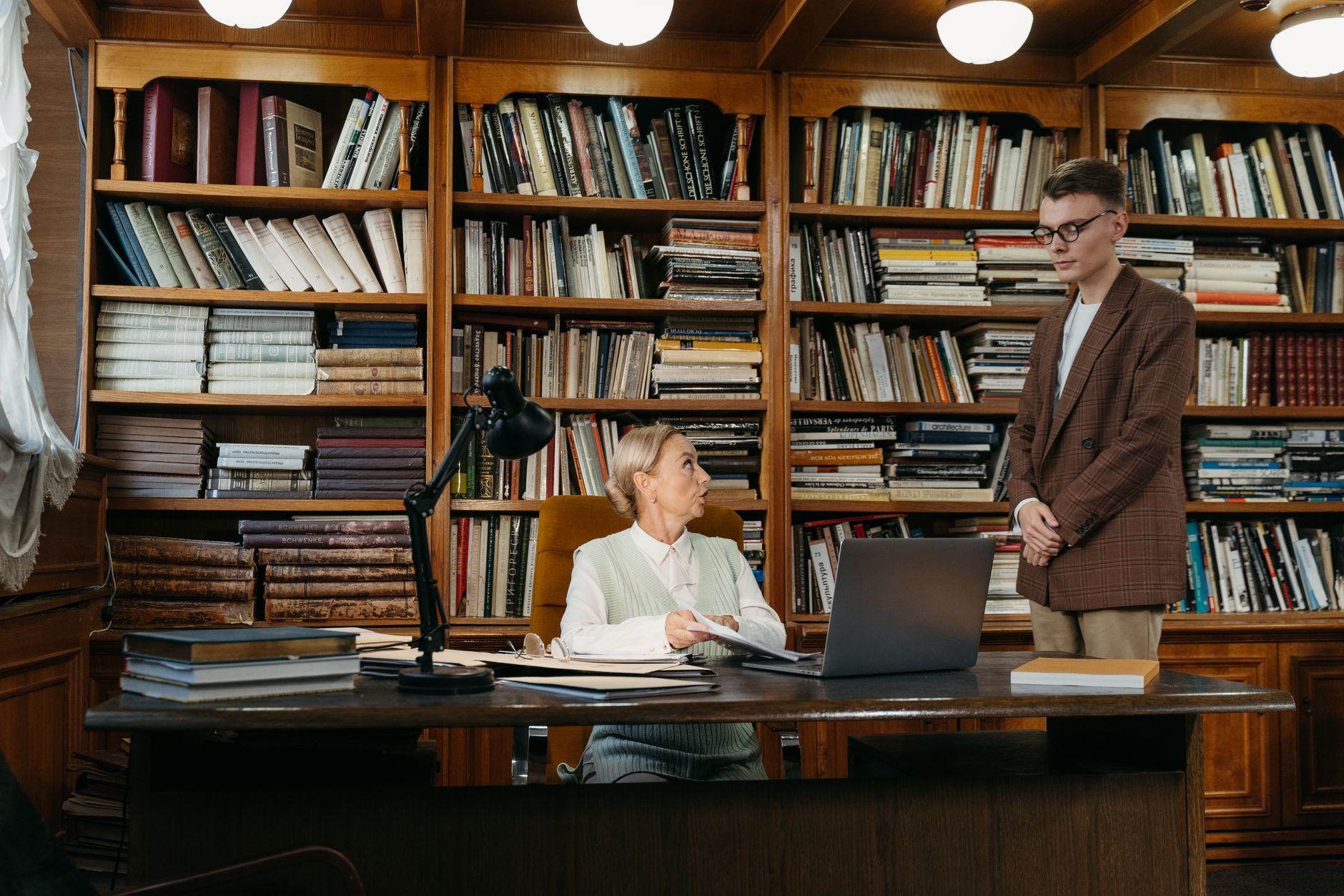 Two people speak across a desk in a library lined with books.